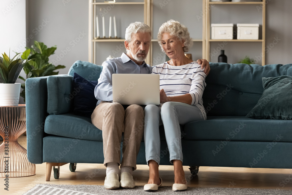 Mature man and woman using laptop together, looking at screen, older spouses browsing apps, shopping or chatting online, making video call, using bank service, sitting on cozy sofa in living room Mature man and woman using laptop together, looking at screen, older spouses browsing apps, shopping or chatting online, making video call, using bank service, sitting on cozy sofa in living room