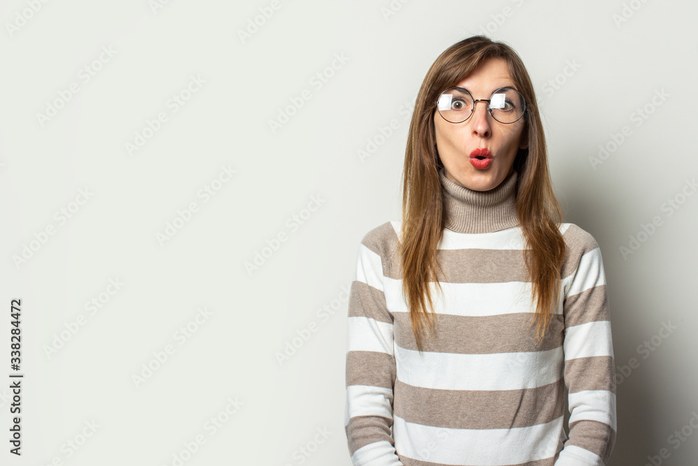 Portrait of a young friendly woman in a striped sweater and glasses with a surprised face on an isolated light background. Emotional face. Gesture surprise shock Portrait of a young friendly woman in a striped sweater and glasses with a surprised face on an isolated light background. Emotional face. Gesture surprise shock