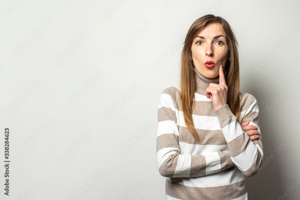Friendly young woman in a striped sweater holding her finger to her chin with thoughtful face on an isolated light background. Emotional face. Gesture to think about something, to dream, to plan Friendly young woman in a striped sweater holding her finger to her chin with thoughtful face on an isolated light background. Emotional face. Gesture to think about something, to dream, to plan
