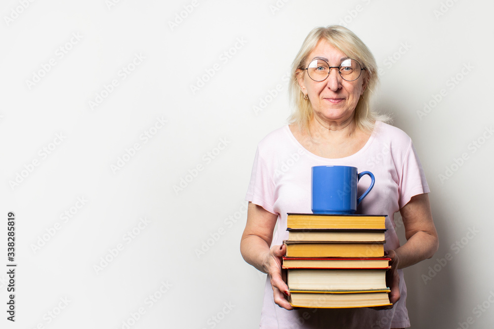 Portrait of an old friendly woman with a smile in a casual t-shirt and glasses holds a stack of books and a cup on an isolated light background. Emotional face. Concept book club, leisure, education Portrait of an old friendly woman with a smile in a casual t-shirt and glasses holds a stack of books and a cup on an isolated light background. Emotional face. Concept book club, leisure, education