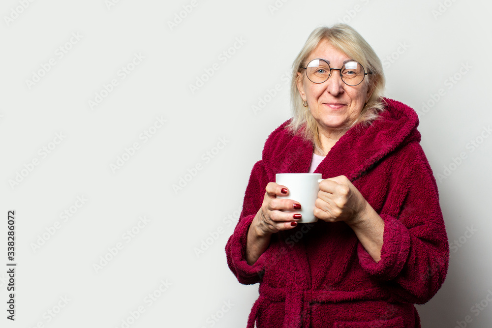 Portrait of an old friendly woman in a casual dressing gown and glasses holding a cup on an isolated light background. Emotional face. Morning coffee concept Portrait of an old friendly woman in a casual dressing gown and glasses holding a cup on an isolated light background. Emotional face. Morning coffee concept