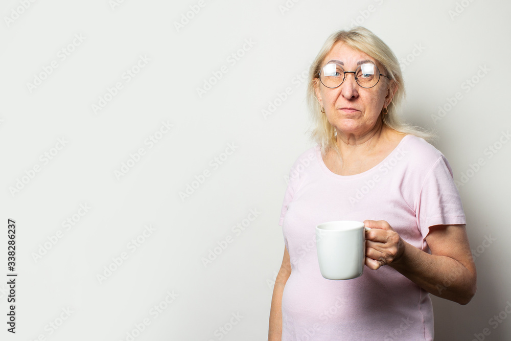 Portrait of an old friendly woman in casual t-shirt and glasses holding a mug in her hands on an isolated white background. Emotional face. Morning coffee concept, breakfast Portrait of an old friendly woman in casual t-shirt and glasses holding a mug in her hands on an isolated white background. Emotional face. Morning coffee concept, breakfast