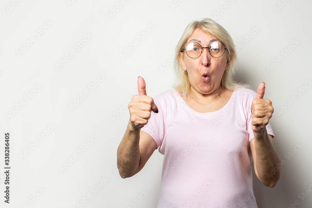 Portrait of an old friendly woman with a surprised face in a casual t-shirt and glasses makes a thumb up gesture on an isolated light background. Emotional face. Gesture is cool, everything is fine Portrait of an old friendly woman with a surprised face in a casual t-shirt and glasses makes a thumb up gesture on an isolated light background. Emotional face. Gesture is cool, everything is fine