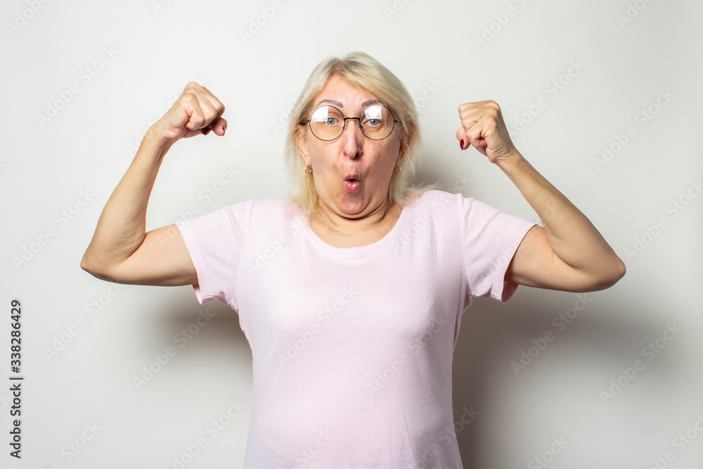 Portrait of an old friendly woman with a surprised face in a casual t-shirt and glasses makes the bodybuilder gesture on an isolated light background. The concept of good health, power Portrait of an old friendly woman with a surprised face in a casual t-shirt and glasses makes the bodybuilder gesture on an isolated light background. The concept of good health, power