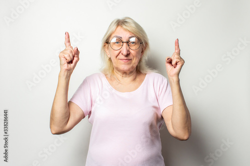 Portrait of an old friendly woman with a smile in a casual t-shirt and glasses points a finger upwards on an isolated light background. Emotional face. Gesture is cool  everything is fine
