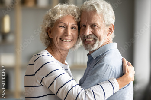 Head shot portrait happy older couple hugging, looking at camera, loving caring mature wife and husband with healthy toothy smiles posing for family photo at home, standing and embracing