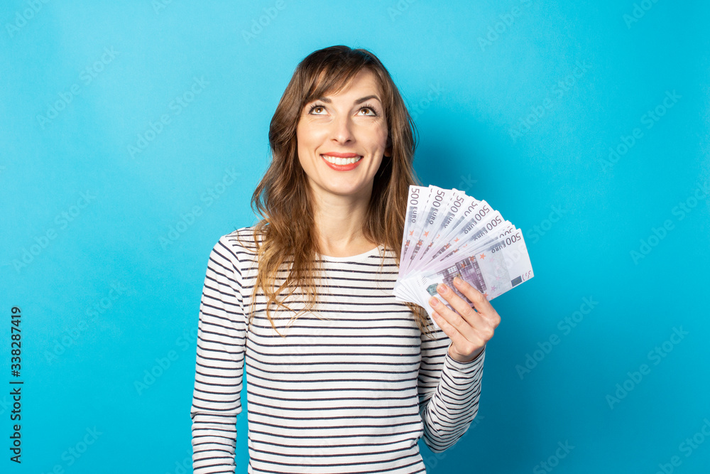 Young friendly woman with a smile in a casual t-shirt holding money in her hands and looking up on an isolated blue background. Emotional face. Concept of wealth, win, credit. Planning, dream Young friendly woman with a smile in a casual t-shirt holding money in her hands and looking up on an isolated blue background. Emotional face. Concept of wealth, win, credit. Planning, dream