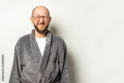 Portrait of a young bald man with a beard in a dressing gown and glasses closed his eyes, squinted at an isolated light background. Emotional face © Alex