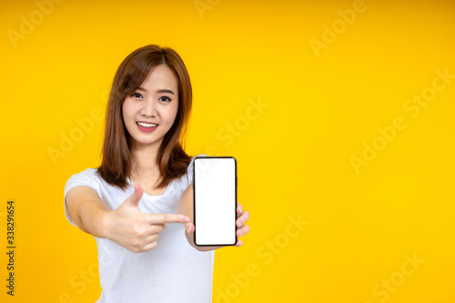 Smiling young Asian woman is pointing on smartphone standing on yellow background.