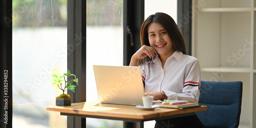 Attractive woman smiling and keep hand on chin while sitting in front her computer laptop at the wooden working desk over comfortable living room as background. © Prathankarnpap