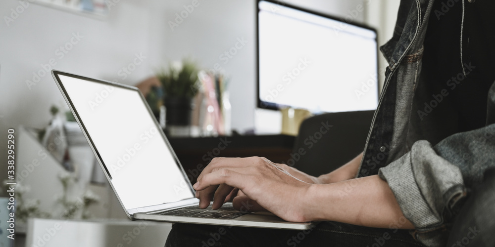 Cropped image of smart man's hands typing on white blank screen computer laptop that putting on his lap over comfortable sitting room as background. Work from home and remote working concept. Cropped image of smart man's hands typing on white blank screen computer laptop that putting on his lap over comfortable sitting room as background. Work from home and remote working concept.