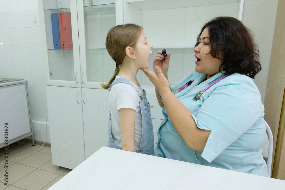 The girl came to the pediatrician with an otoscope. Smiling otolaryngologist doctor looking at a child while examining a child's throat with a tongue depressor. The girl came to the pediatrician with an otoscope. Smiling otolaryngologist doctor looking at a child while examining a child's throat with a tongue depressor.