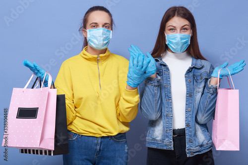 Closeup portrait of two young girls wearing casual outfits and protective masks and gloves, holding shopping bags, spreading hands aside, con not resist shopping during . Coronavirus, Civid19 concept. photo