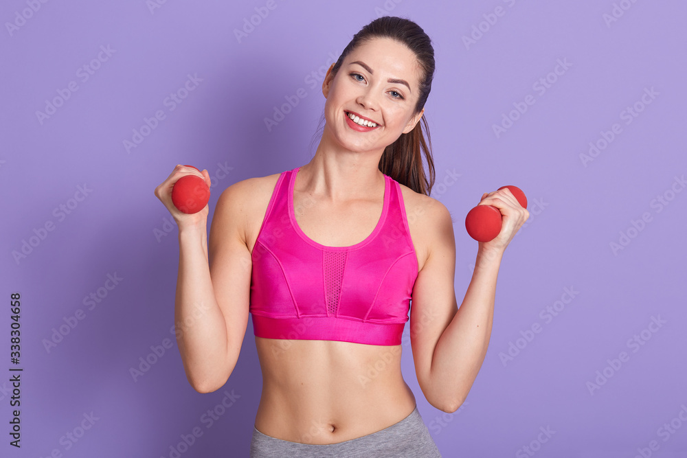 Horizontal portrait of cheerful positive fitness trainer standing isolated over lilac background in studio, holding dumbbells, looking directly at camera, having training, keeping fit. Sport concept. Horizontal portrait of cheerful positive fitness trainer standing isolated over lilac background in studio, holding dumbbells, looking directly at camera, having training, keeping fit. Sport concept.