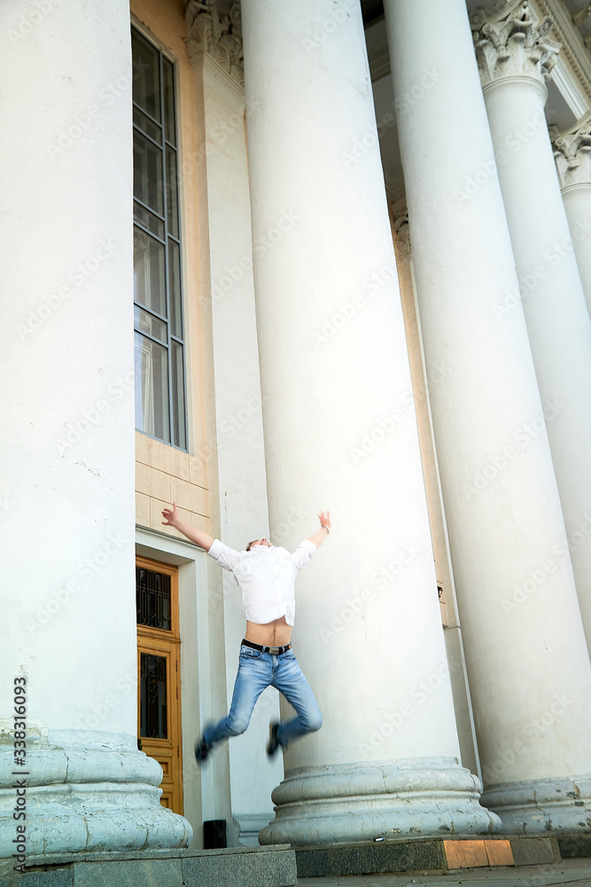 Young handsome man in jeans and a white shirt jumping near building with white columns