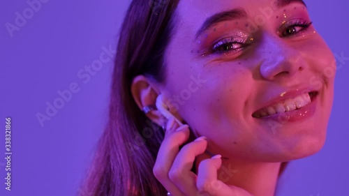 A stylish woman with fashion glitter makeup is turning on her wireless earphones isolated over violet neon background