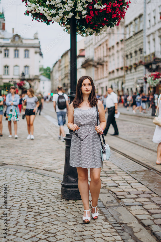 Beautiful girl in a summer dress walks the city. Brunette girl in a sundress with a handbag and glasses. Girl on the background of the city