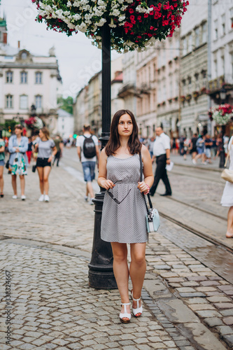 Beautiful girl in a summer dress walks the city. Brunette girl in a sundress with a handbag and glasses. Girl on the background of the city