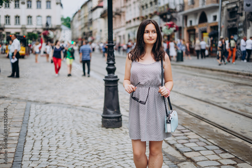 Beautiful girl in a summer dress walks the city. Brunette girl in a sundress with a handbag and glasses. Girl on the background of the city Beautiful girl in a summer dress walks the city. Brunette girl in a sundress with a handbag and glasses. Girl on the background of the city