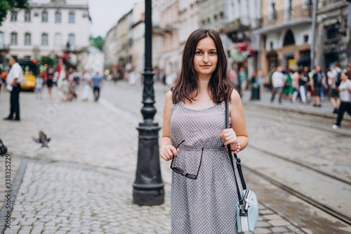 Beautiful young woman walking through the old city. Brunette girl in dress with handbag and glasses. Woman and city in summer sunny day.