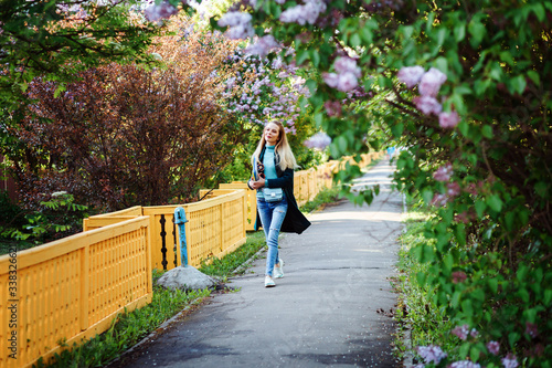 Attractive woman is walking through the lilac alley in spring. Student girl is spending time in the countryside. 