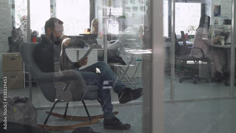 Modern international office, man is sitting in a rocking chair and working on a tablet and drink coffee, office workers and managers working at computers with documents, view through the glass. Modern international office, man is sitting in a rocking chair and working on a tablet and drink coffee, office workers and managers working at computers with documents, view through the glass.