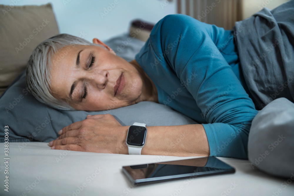 Fototapeta premium Sleep Apps - Tech-savvy Senior Woman Sleeping in Bed, Using Smart Phone and Smart Watch to Improve her Sleeping Habits