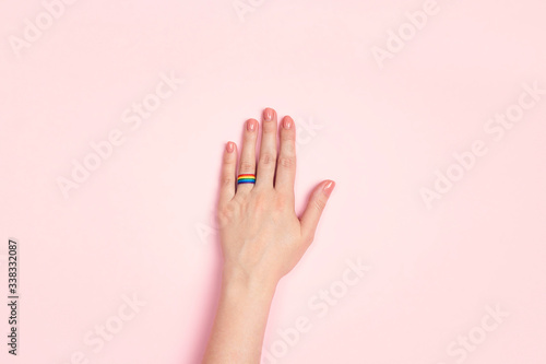Female hand with a wedding ring from a rainbow ribbon on a pink background. Lesbian marriage proposal.