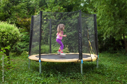 Garden trampoline with little child moving inside. Trampolin in the garth with little girl jumping. Fun on garden trampoline in summer day. Recreational trampoline with a safety net enclosure. photo