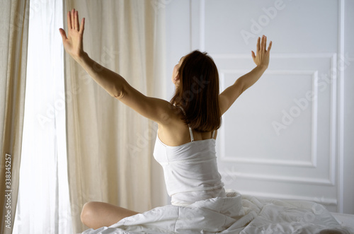 Woman stretching in bed after waking up, back view, entering a day happy and relaxed after good night sleep. Good morning and new day for brunette. Concept of Weekend, holidays