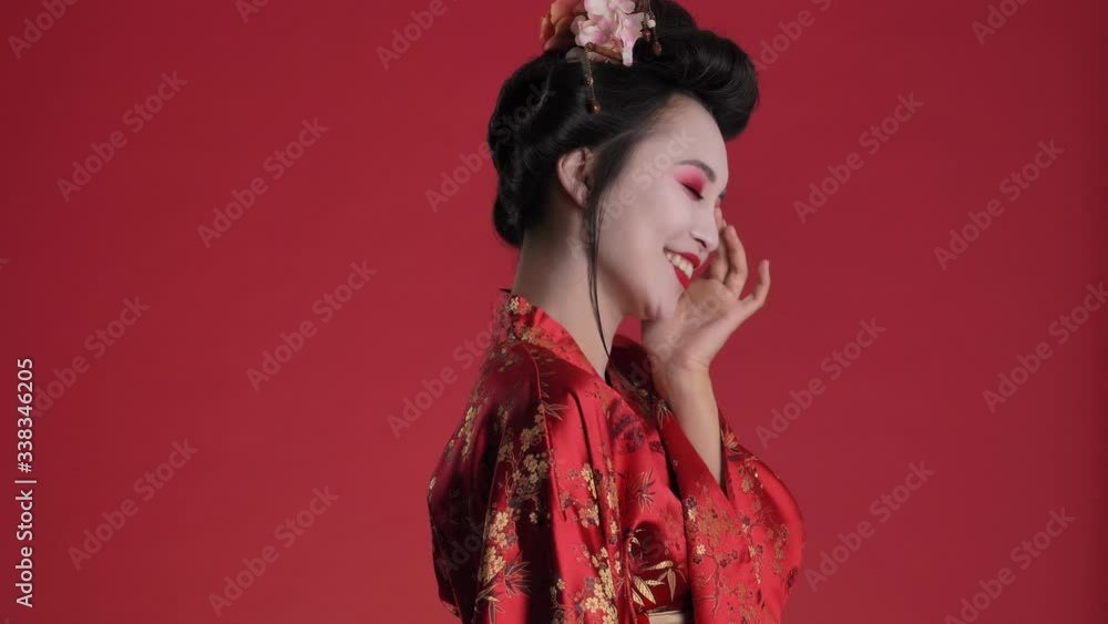 A pretty nice young geisha woman in traditional japanese kimono is posing while turning to the camera isolated over red background A pretty nice young geisha woman in traditional japanese kimono is posing while turning to the camera isolated over red background