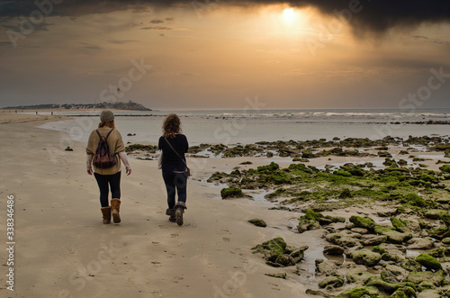 Back view of two women walking on a beach of Cadiz called Zahora.  Selective focus on the rocks covered with moss. Trafalgar lighthouse and beautiful sunset in the background. photo