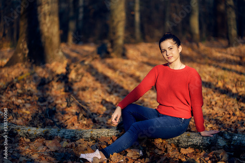 young woman in a red sweater and jeans sits on a log in nature in the autumn forest with fallen golden leaves © Aleksandr