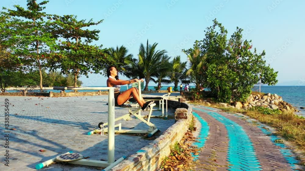 Attractive woman in sportswear exercising on outdoor gym of resort with beautiful garden in front of blue sea in Indonesia Attractive woman in sportswear exercising on outdoor gym of resort with beautiful garden in front of blue sea in Indonesia