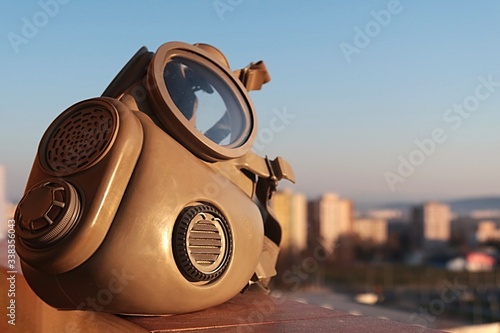 M10 Military grade gas mask with inhale and exhale valves visible, sunbathing in morning sunshine, cityscape in background. 