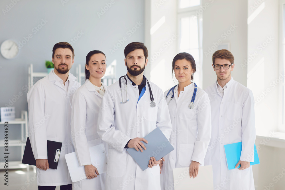 A group of confident practicing doctors in white coats are smiling against the backdrop of the clinic. A group of confident practicing doctors in white coats are smiling against the backdrop of the clinic.