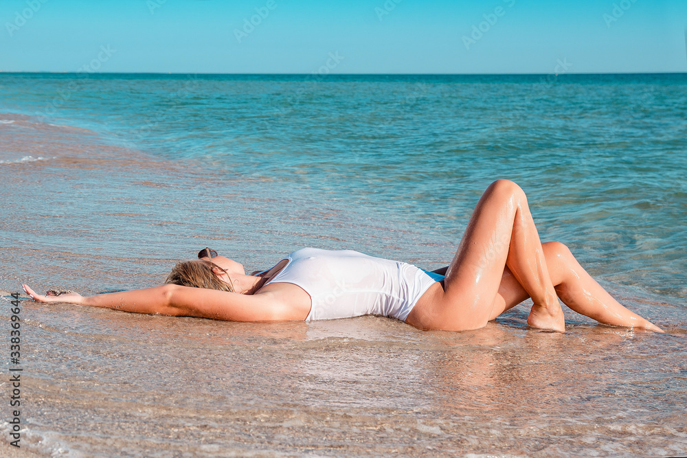 Beautiful girl sunbathing on a sandy tropical beach. Fashion young female lying on back on sand with white swimsuit. Tanned blonde woman relaxing Beautiful girl sunbathing on a sandy tropical beach. Fashion young female lying on back on sand with white swimsuit. Tanned blonde woman relaxing