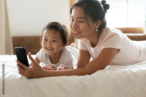 Asian mum and little daughter in pyjamas after awakening have fun use cellphone watch cartoons, mom show kid girl educational on-line program. Parental control, modern tech, younger generation concept