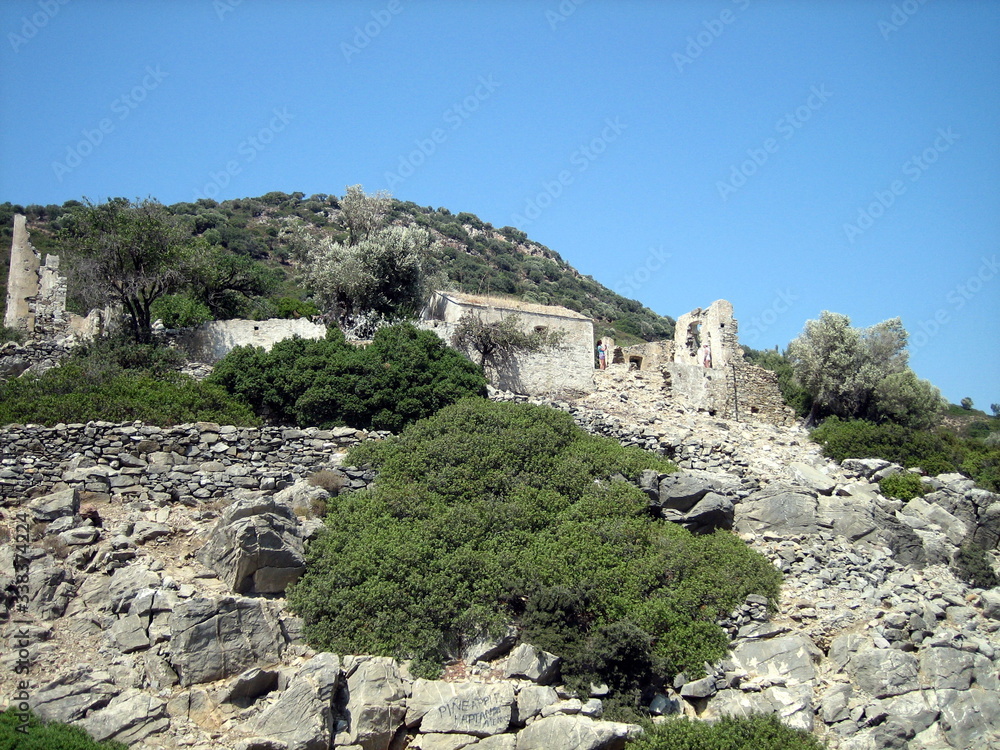 Stone ruins of an ancient building on the slope of a rocky hill, overgrown with green dense vegetation, in good weather.