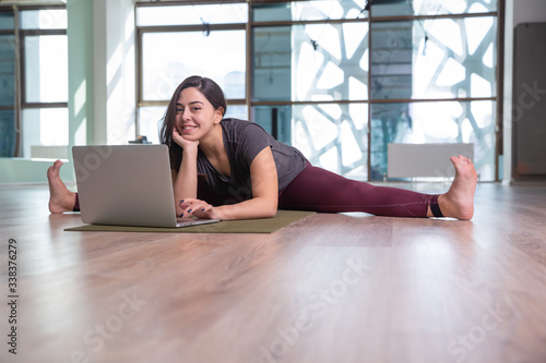 Photo of young woman practicing yoga with laptop indoor. Beautiful girl practice yoga in class. Yoga studio instructor. Blurred background.