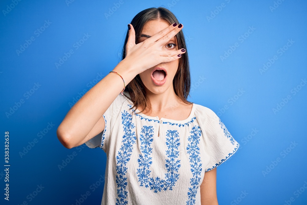 Young beautiful brunette woman wearing casual t-shirt standing over blue background peeking in shock covering face and eyes with hand, looking through fingers with embarrassed expression. Young beautiful brunette woman wearing casual t-shirt standing over blue background peeking in shock covering face and eyes with hand, looking through fingers with embarrassed expression.