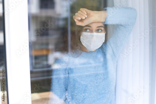 Young woman in quarantine wearing a mask and looking through the window photo