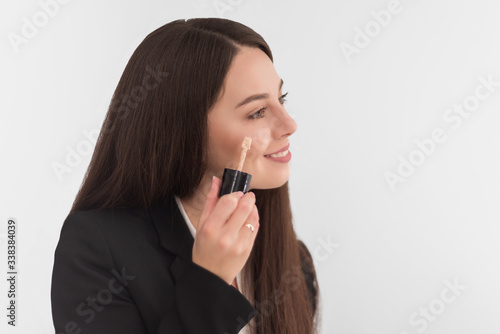 girl, make-up artist, with long dark hair in a business suit, applies a concealer on the face on a white background