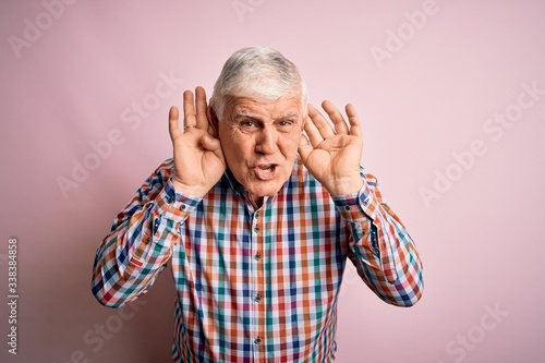 Senior handsome hoary man wearing casual colorful shirt over isolated pink background Trying to hear both hands on ear gesture, curious for gossip. Hearing problem, deaf