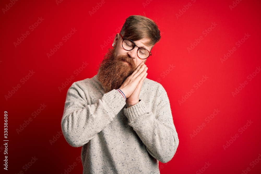 Handsome Irish redhead man with beard wearing casual sweater and glasses over red background sleeping tired dreaming and posing with hands together while smiling with closed eyes. Handsome Irish redhead man with beard wearing casual sweater and glasses over red background sleeping tired dreaming and posing with hands together while smiling with closed eyes.