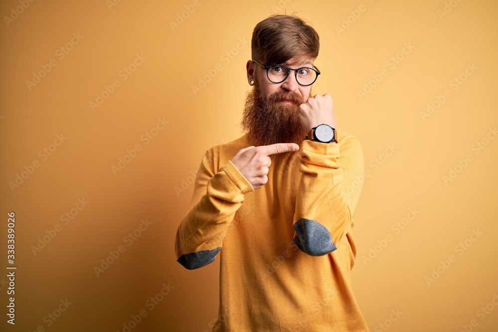 Fototapeta premium Handsome Irish redhead man with beard wearing glasses over yellow isolated background In hurry pointing to watch time, impatience, looking at the camera with relaxed expression