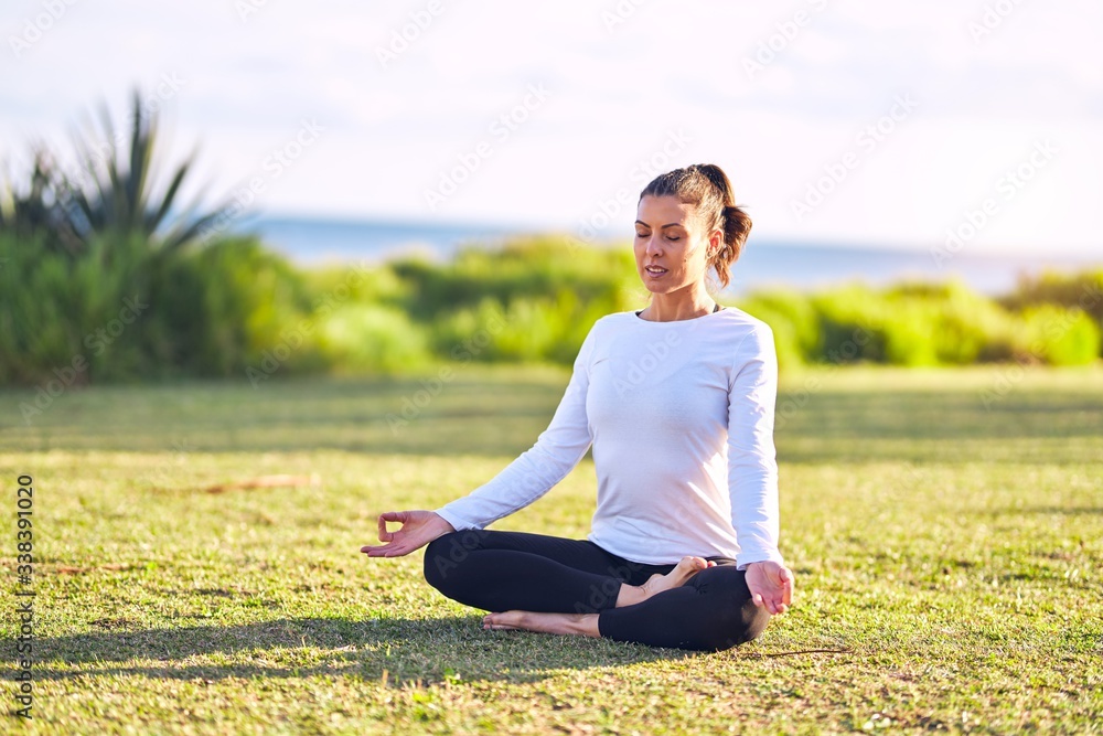 Young beautiful sportwoman practicing yoga. Coach sitting on the grass doing lotus pose at park Young beautiful sportwoman practicing yoga. Coach sitting on the grass doing lotus pose at park