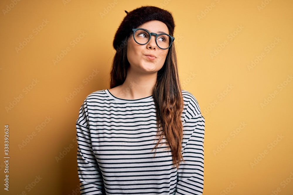 Young beautiful brunette woman wearing french beret and glasses over yellow background smiling looking to the side and staring away thinking. Young beautiful brunette woman wearing french beret and glasses over yellow background smiling looking to the side and staring away thinking.