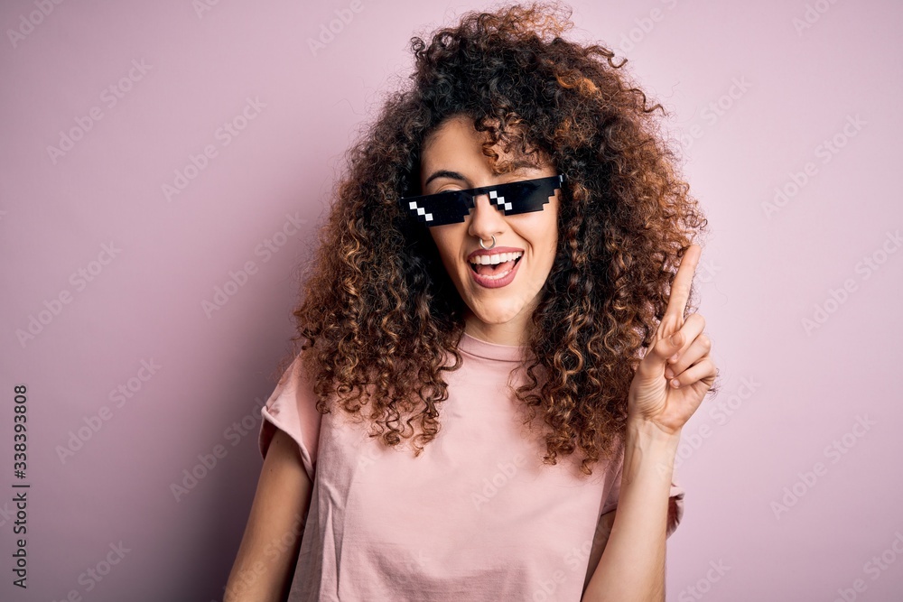 Young beautiful woman with curly hair and piercing wearing funny thug life sunglasses pointing finger up with successful idea. Exited and happy. Number one.