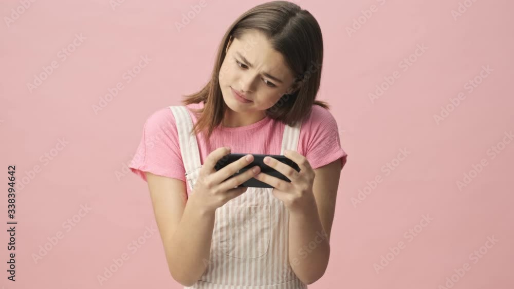 Upset pretty brunette woman in overalls playing on smartphone over pink background Upset pretty brunette woman in overalls playing on smartphone over pink background